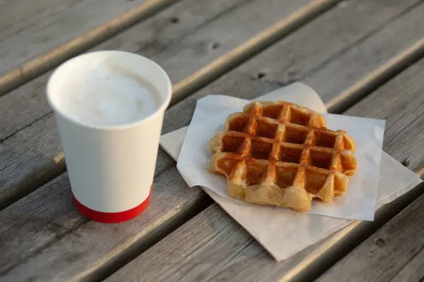 Kağıt bardakta kırmızı şeritli bir kapuçino ve açık havada tahta bir masada Belçika waffle 'ı..