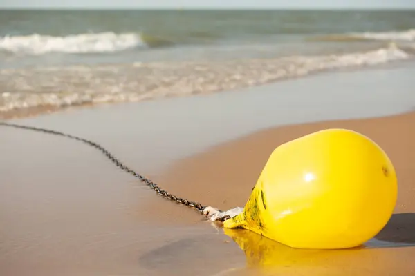 Paslı zincirli sarı şamandıra. Plastik atıkla bağlanmış. Islak kumsalda güneş ışığı altında yatıyor. Plastik kirliliği, insan atıkları ve deniz hasarının sembolü