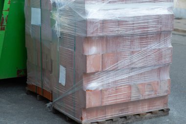 İnşaat alanındaki ahşap bir paletin üzerinde duran şeffaf plastik germe filmine sıkıca sarılı kırmızı yapı tuğlaları yığını. Yenileme, endüstriyel malzemeler ve kentsel gelişim kavramı.