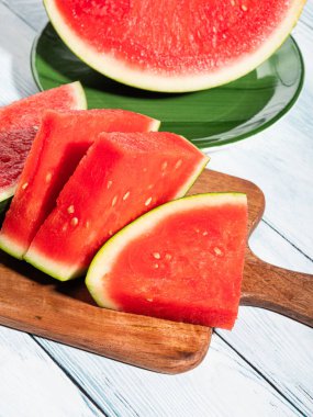 Köy arka planında tahta kesme tahtasındaki çekirdeksiz karpuz dilimleri sert ışıkla aydınlatılıyor. Taze meyveli canlı yaz sahnesi, yakın çekim.