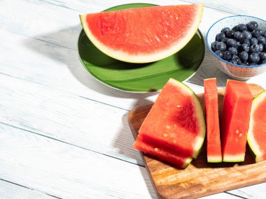 Sert ışıkla aydınlatılmış taze kesilmiş çekirdeksiz karpuz dilimleri ve rustik ahşap arka planda yabanmersini. Taze meyveli canlı yaz sahnesi, kopyalama alanı