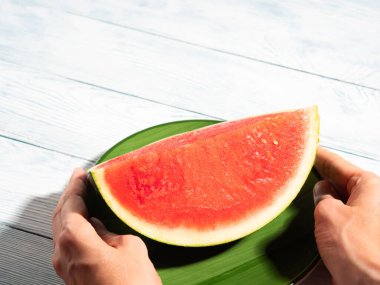 Yeşil tabakta tohumsuz karpuz. Kırsal arka planda sert ışıkla tutulan dişi eller tarafından tutulmuş. Taze meyveli canlı yaz sahnesi