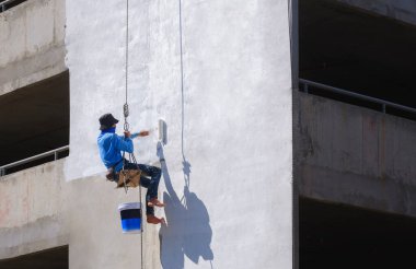 İnşaat alanındaki otopark binasının dış duvarındaki beyaz astar boyaya iple inen Asyalı inşaat işçisi