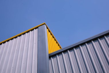 Modern depo binasının gri ve sarı kıvrımlı çelik duvarının alttaki görüntüsü mavi açık gökyüzü arka planına karşıdır.