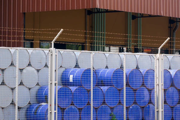 Many blue and white 200 liter chemical or oil drum stacking behind mesh fence inside of industrial factory area