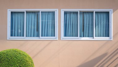 Row of glass sliding windows and blue sunshade on beige gypsum board wall with round shrub on foreground, home exterior architecture concept