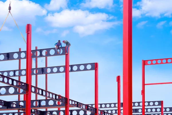 2 construction workers with crane are installing black castellated beam on red metal poles of factory building structure in construction site area with blue sky