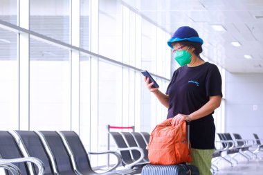Havaalanı terminalindeki yolcu dinlenme yerinde dururken akıllı telefon kullanan koruyucu maskeli Asyalı kadın turist.