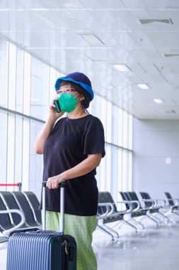 Yüklü koruyucu maske takan Asyalı kadın yolcu havaalanındaki dinlenme yerinde dikey karede beklerken cep telefonuyla konuşuyor.