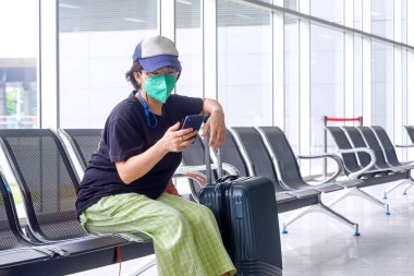 Koruyucu maskeli Asyalı kadın turist havaalanı terminalinde yolcu dinlenme alanında yolcu beklerken bankta akıllı telefon kullanıyor.