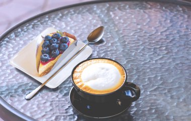 Sütlü kahve fincanında sıcak latte kahve, yaban mersinli turta ve yuvarlak cam sehpada kaşık.