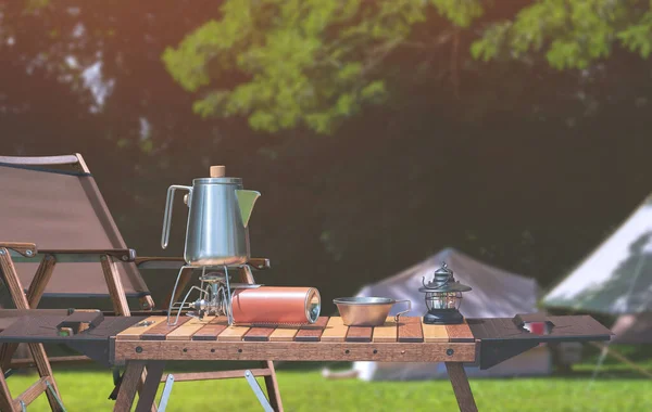 Açık hava kamp masası, paslanmaz çelik çaydanlık, gaz sobası ve kamp alanındaki yeşil çimlerin üzerine eski model fener.