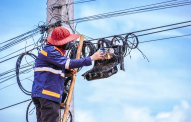 Ahşap merdivendeki teknisyen, elektrik direğindeki internet bağlantı sistemini mavi gökyüzü arkaplanıyla karşılaştırırken ayırıcı kutusundaki fiber optik kabloları kontrol ediyor.