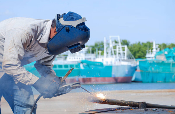 Welder with safety equipment using arc welding machine to welding galvanized steel pipe in shipyard area at harbor