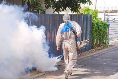 Rear view of healthcare worker in protective clothing using fogging machine spraying chemical to eliminate mosquitoes on street in alley of public suburb community clipart