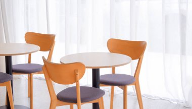 Modern kahve dükkanının beyaz perdesinin yanında bej renkli iki ahşap yemek masası.