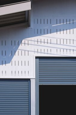 İki tekerlekli panjurlu giriş kapısı, beyaz havalandırmalı çimento. Depo binasının duvarında güneş ışığı ve gölgesi dikey çerçevede.