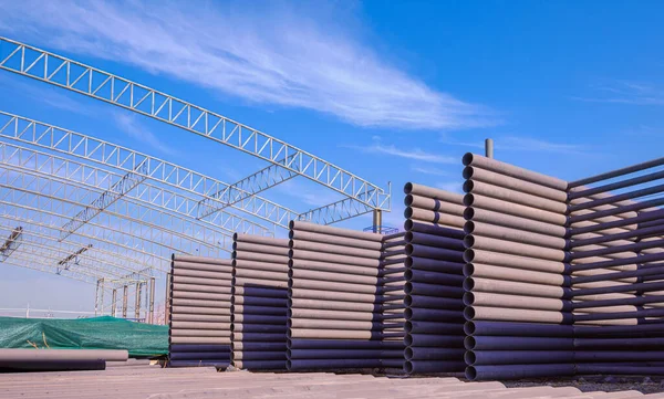 Piles of many carbon steel roof beams for installing on top of large industrial factory building structure in construction site area against blue sky background