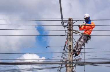 Güvenlik ekipmanı ve çeşitli iş araçlarına sahip elektrikçi. Elektrik direğine kablo hatları ve elektrik sistemi kurarak bulutlu gökyüzü arka planına karşı