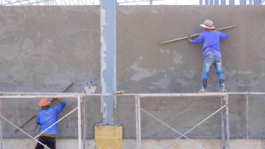 İnşaat alanındaki inşaat alanının beton duvarına çimento yapıştırmak için uzun malalar kullanan iki inşaat işçisi.