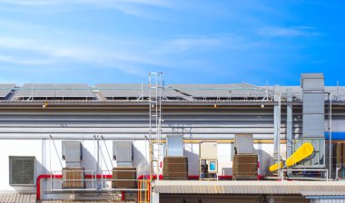 Exhaust duct and ventilation equipment in EVAP system on the wall with solar panels on steel roof of industrial building against blue sky background clipart