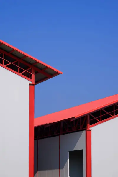 Part of 2 industrial cold storage warehouse buildings with door frame on white sandwich panel wall and red metal gable roof in construction site against blue sky background in vertical frame