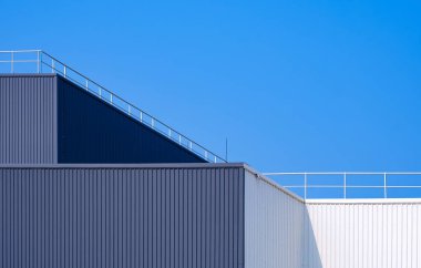 Gri ve beyaz alüminyum kıvrımlı fabrika binası geometrik minimal tarzda, çatısı çelik çitle çevrili, mavi gökyüzü zeminine karşı.