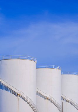 Petrol endüstrisi bölgesinde üç beyaz yakıt deposu dikey çerçevede mavi gökyüzü arkaplanına karşı