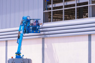 Eklemli asansördeki inşaat işçisi inşaat alanındaki sanayi fabrikası iskeletine alüminyum kıvrımlı çelik levhalar yerleştiriyor.