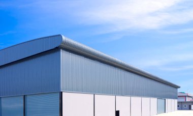 Fabrika alanındaki beton ve kıvrımlı alüminyum duvarlı patenli kapılı büyük bir sanayi deposu. Mavi gökyüzü arka planı, düşük açı ve yan görüş açısı.
