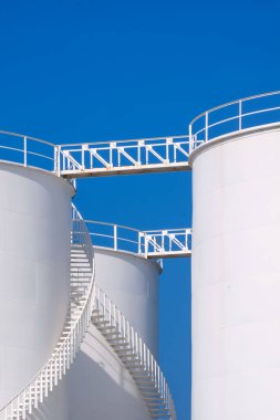 Beyaz depo yakıt tankları dikey çerçevede mavi berrak gökyüzü arkaplanına karşı sarmal merdiven, düşük açı görünümü 