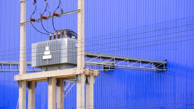 Elektrik trafosu ve elektrik direğinde yüksek voltaj dağıtım ekipmanı. Tepedeki kablo merdiven tepsisi mavi alüminyum kıvrımlı sanayi fabrikası binasının metal duvarının yanında.