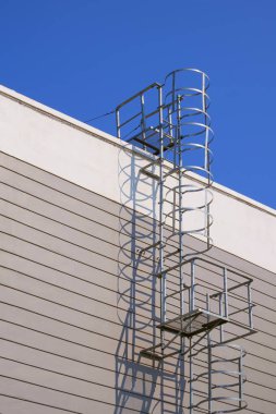 Cylindrical ladder on gray and white warehouse industrial building wall against blue sky background, low angle view and vertical frame