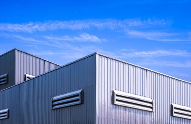 Endüstriyel depo fabrika binası, gri alüminyum kaplamalı çelik duvar, mavi gökyüzü arka planına karşı, modern tarzda geometrik dış sanayi mimarisi.