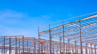 Geometrik metal sütunlar ve iki depo fabrikasının çatı kiriş yapısı büyük inşaat sahasında mavi gökyüzü arka planına karşı, yan görüş