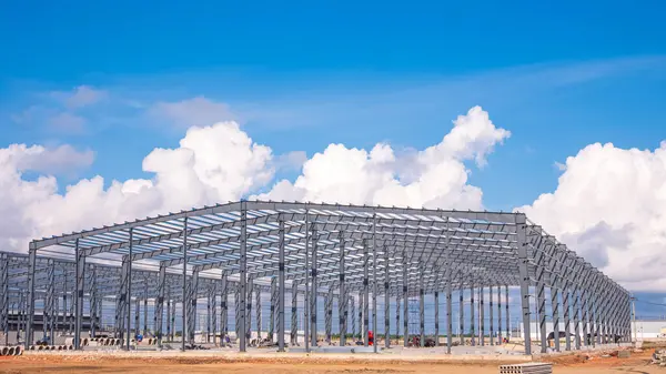 İnşaat alanındaki fabrika deposunun büyük metal yapısı mavi gökyüzü arka planındaki bulutlara karşı.
