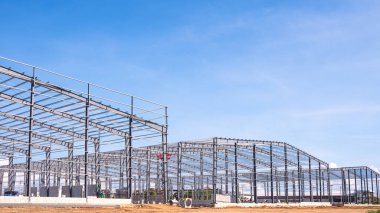 Üç endüstriyel depo fabrika binasının metal sütunları ve çatı kiriş yapısı işçilerle birlikte büyük inşaat sahasında mavi gökyüzü arka planına ve yan manzaraya karşı çalışıyor