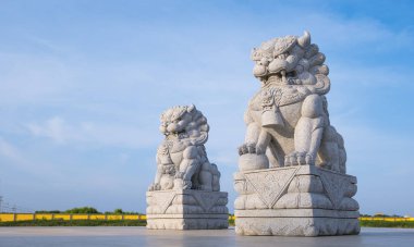 Two carved stone Chinese lion statues on concrete yard floor against blue sky background, perspective side view with copy space