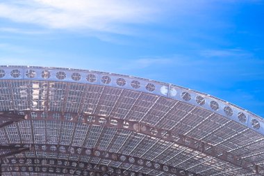 Eğri çatı kirişlerinin tepesindeki birçok güneş paneli, mavi gökyüzü arkaplanına karşı metal hücresel ışınlardan yapılmış yapı, düşük açı ve kopyalama alanı