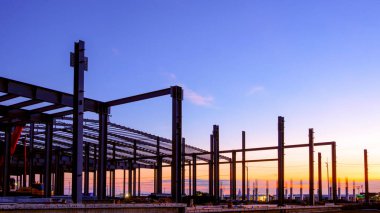 İnşaat alanında metal sütunları ve çatı kiriş iskeleti olan siluet endüstriyel yapı, renkli gökyüzü arka planına karşı, perspektif yan görünümü