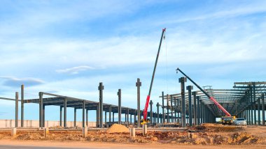 Fabrika endüstriyel yapı iskeleti işçilere ve vinç kamyonlarına ait metal çatı kirişini mavi gökyüzü arka planına karşı inşaat alanındaki depo yapısının tepesine yerleştirmek için kaldırıyor.