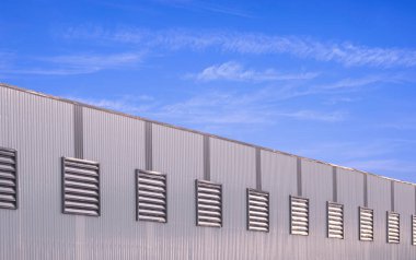 Büyük endüstriyel fabrika üretim binası. Gri alüminyumun üzerinde sıra sıra havalandırma lopları var. Mavi gökyüzü arka planına karşı çelik duvar.