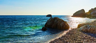 Berrak turkuvaz suyu, güneş ışığı dalgaları ve su altındaki taşlarla kayalık bir sahil manzarası parlak bir gökyüzü altında canlı bir kıyı manzarası yaratıyor..