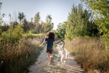 Bir kız ormanda toprak bir yolda beyaz bir köpekle koşuyor. Tasmalı bir köpek. Köpek tasmayı ısırır..