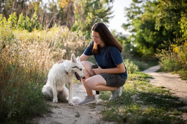 Bir kız ormandaki toprak yolda beyaz bir köpeği okşuyor..
