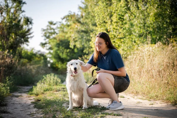 Kız ormanın arka planında beyaz bir köpeği okşuyor. Toprak bir yoldalar..