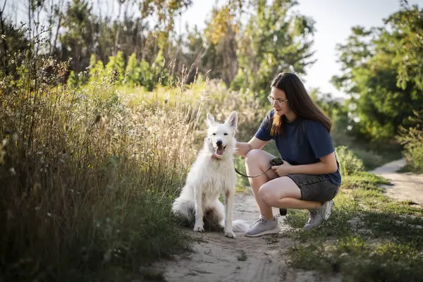 Gözlüklü bir kız ormanda toprak bir yolda beyaz bir köpeği okşuyor..