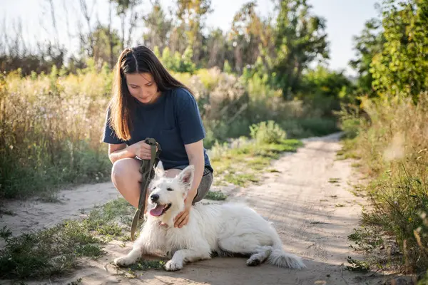 Bir kız, gündüz vakti bir ormanın arka planında toprak bir yolda yatan bir köpeği okşar..