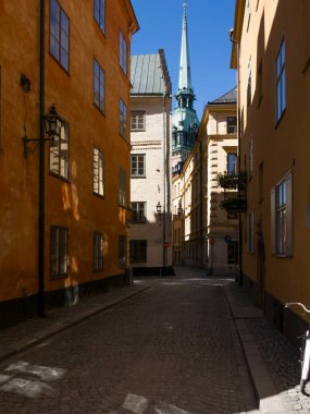 Stockholm, İsveç 'in tarihi eski kasabasında (Gamla Stan olarak bilinir) uzun, boyalı eski ve tarihi binalarla kaplı dar bir kaldırım taşı caddesi. Arka planda mavi gökyüzü içinde yeşil bir kilise kulesi var..