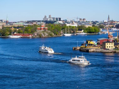 İki beyaz feribot İsveç 'in Stockholm limanı yakınlarındaki bir su yolundan geçiyor. Arka planda, verimli yeşillik, kıyı boyunca uzanan eski ve yeni binalar, vinçler ve sanayi önde gelen.